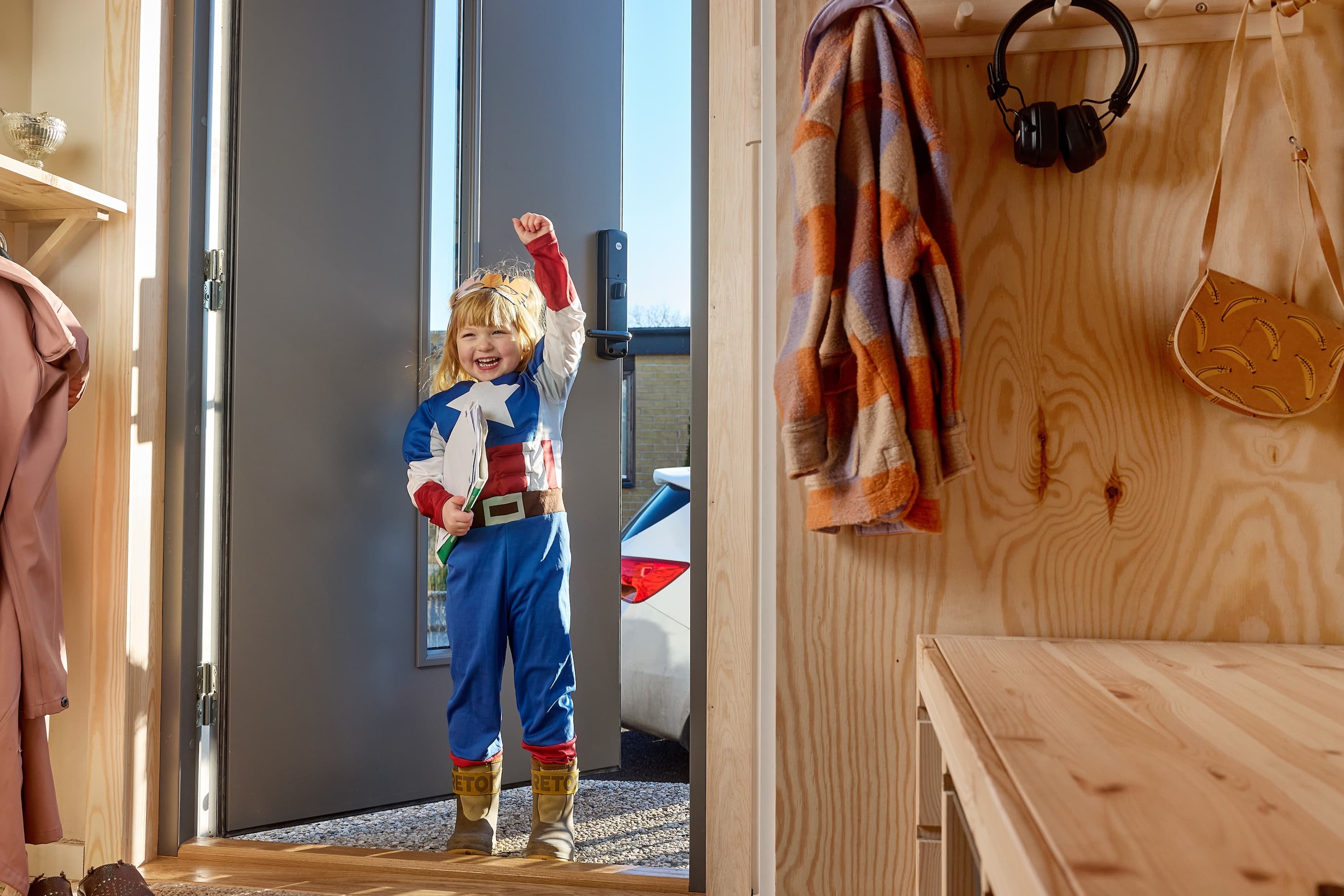 Child in a superhero costume excitedly entering a room with wooden shelves holding clothes, a bag, and headphones.